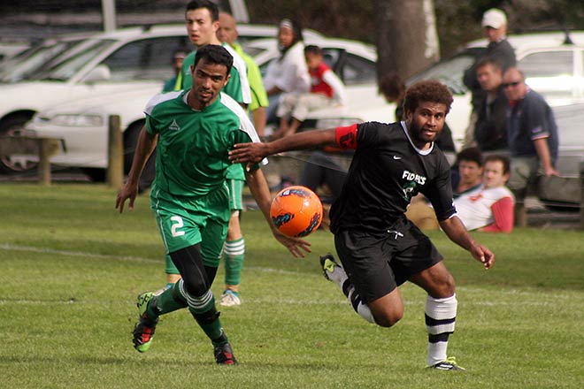 Two men chase an orange football during a football game, wearing different football uniforms. A small crowd watches from the sideline.