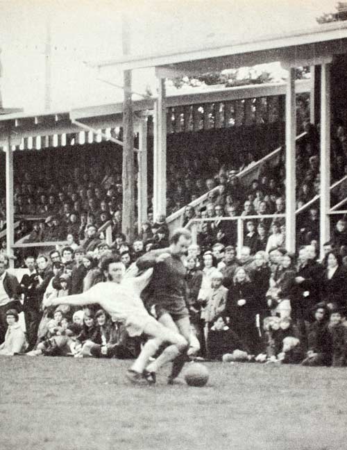 Two men in a football game tussle for a ball at their feet, playing in front of a large crowd seated in a stadium.