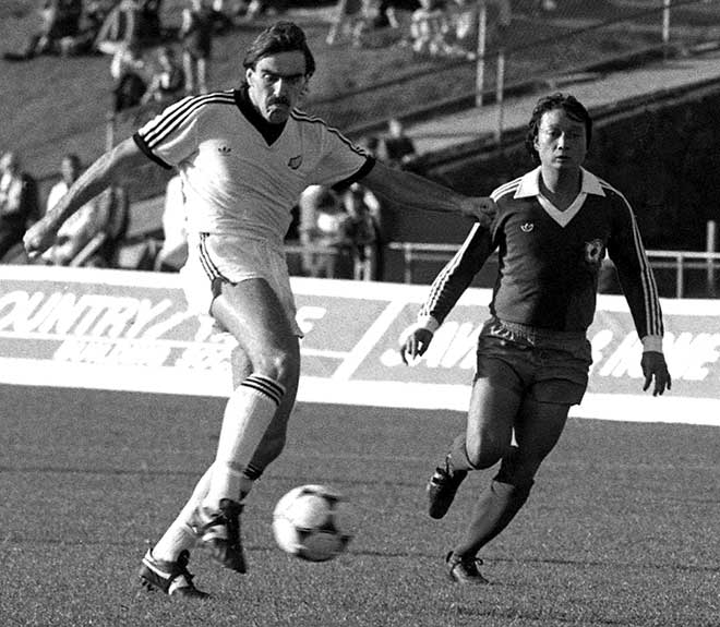 A man in a white New Zealand football uniform kicks a ball while a member of another team chases behind him during a football game.