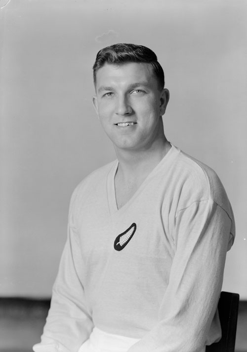 A man seated for a formal portrait wearing a white New Zealand football uniform with a silver fern badge on his uniform.