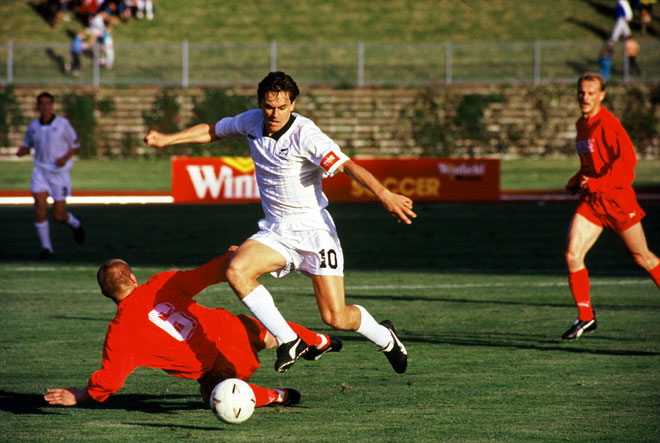 A man in a white New Zealand football uniform leaps in the air to kick a ball during a football game while a man in a red uniform has fallen on the ground. 
