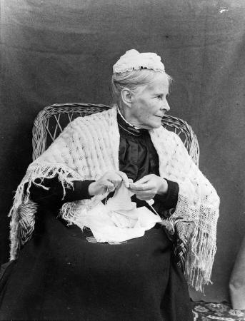 Anna Maria Williams in later life, seated in a wicker chair, wearing a crocheted hat and shawl, and knitting.. 