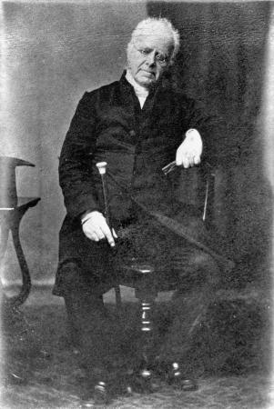 Henry Williams in later life, dressed formally, seated in a chair and holding a cane.