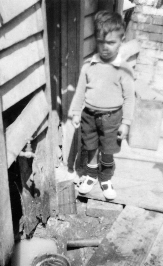 Small boy standing next to an open drain outside the kitchen of a run-down house.