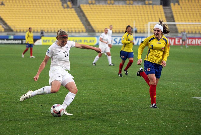 A woman in a white New Zealand football uniform kicks the ball during a football game in a stadium, watched by members of her and the opposing team.