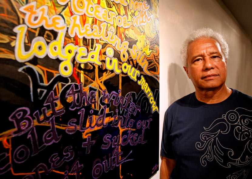 Albert Wendt in black T-Shirt standing next to art work on wall.