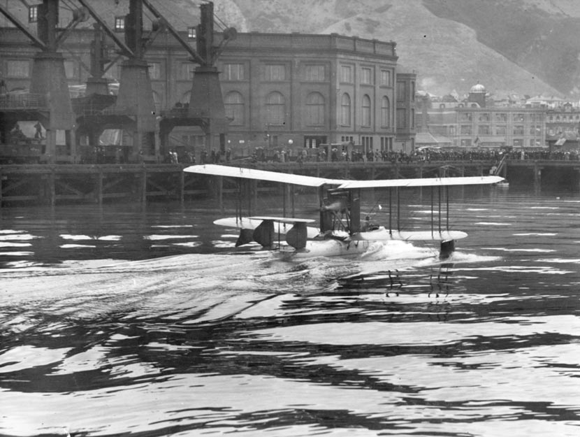 A photograph showing a seaplane moving through water beside the Wellington wharves that are lined with people