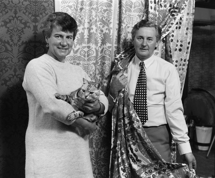A man and woman stand surrounded by textile samples. The woman holds a cat. 