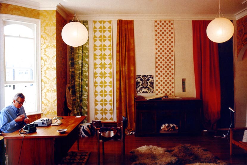 Man seated at a desk in a showroom with textile and wallpaper samples hanging on the walls.