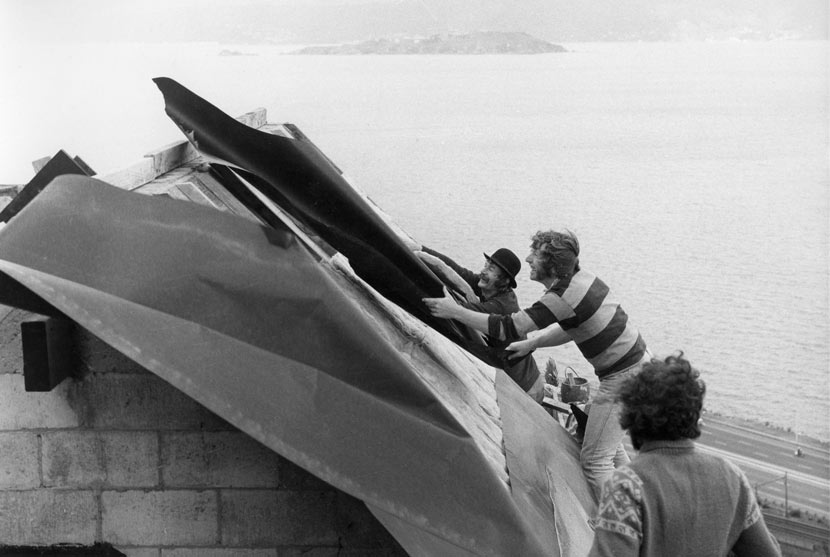 A photograph of three men working on the roof of Athfield House with Wellington Harbour and Somes Island in the background