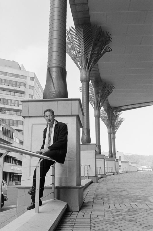 A photograph of Ian Athfield seated at the bottom of a paved ramp with a roof supported by columns sculpted to look like nikau palms