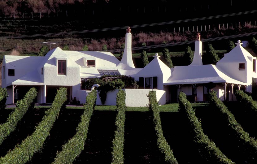 A photograph of a white stucco building with two chimneys with rows of grape vines in the foreground