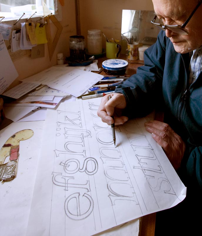 A photograph of Joseph Churchward seated at a desk and sketching a typeface design on a sheet of paper