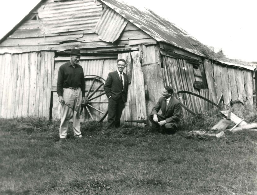 A photograph of three men sitting and standing outside a falling-down historic building built of timber and patched with iron.