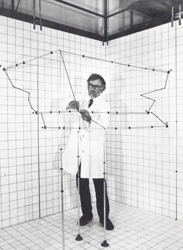 Man stands inside a small room with a grid pattern on the walls and floors, also containing rods and spheres.