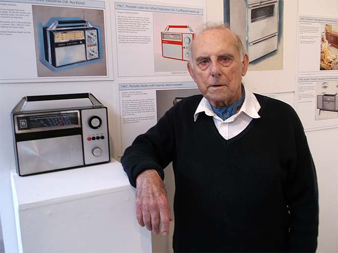 Gifford Jackson beside a radio he designed with illustrations of other designs on the wall behind him