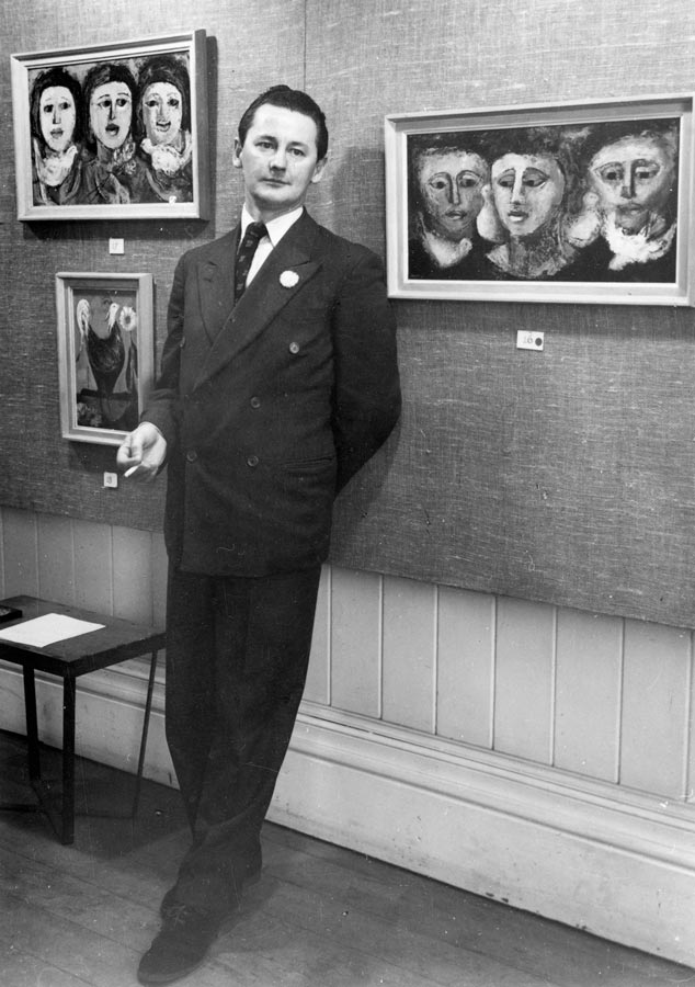 A man in a suit stands in front of painting which are hung on the wall of a gallery.