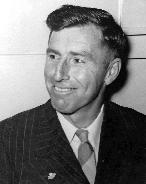 A black and white head and shoulders photograph of Roly Earp, smiling, wearing a suit and tie. 