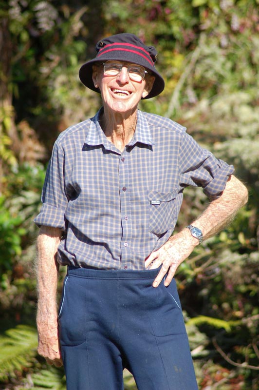 A colour photograph of Roly Earp in old age, standing outdoors in a casual pose and smiling broadly.