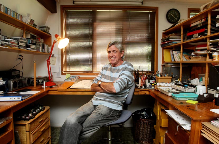 Murray Ball, in later life, is seated at his drawing desk, surround by art supplies, shelving with books and ringbinders, and a tape deck and cassettes. 