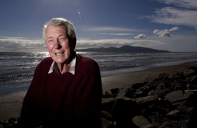 Ian Cross in old age, standing on a beach with the ocean and Kāpiti Island in the background.