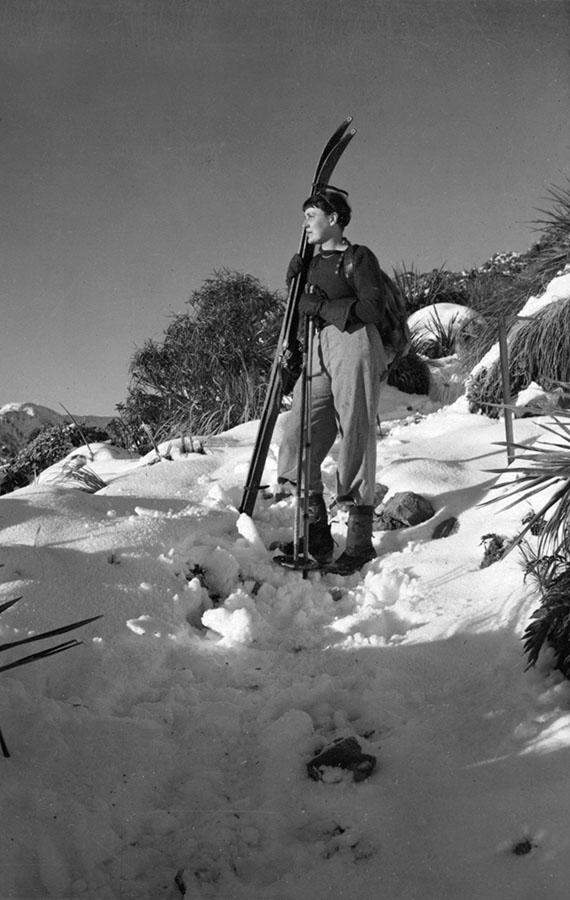 Mavis Davidson as a young woman, standing in profile on a snow-covered slope, holding skis and ski-poles.