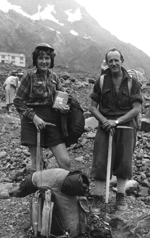 A man and a woman, holding ice-climbing pickaxes, stand on a rocky alpine slope with a mountain and hut in the background. 
