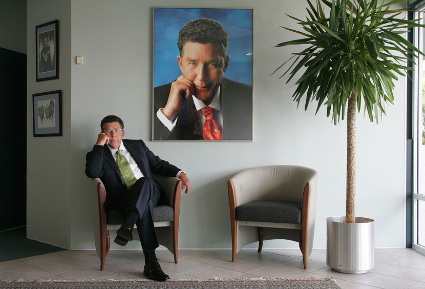 Paul Holmes, seated in a corporate office, resting his head on his hand, mirroring his posture in a large, framed photograph of himself which hangs directly behind him. 