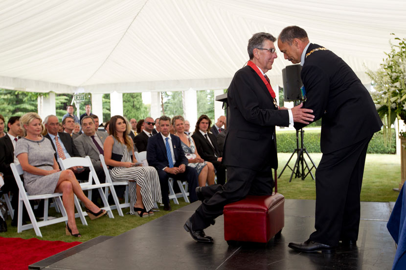 Paul Holmes kneels on a stool, while Jerry Mateparae places a mdeal around his neck. The outdoors ceremony is held under a marquee, with Holmes and Mateparae on a low stage and the audience seated on folding chairs. 