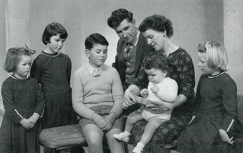 A studio portrait of the Hutchins family, smartly dressed, some seated and some standing. The children are mostly primary school age. All are watching the baby, who plays with a small toy guitar. The three girls wear matching dresses.