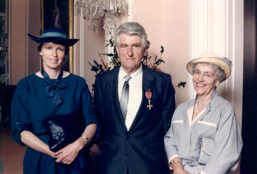 A man and two women, smartly-dressed, standing in front of a chandelier and oil painting. Hutchins wears the OBE pinned to his suit jacket.