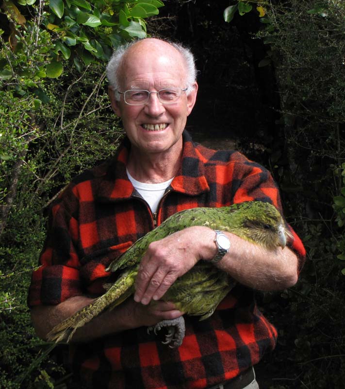 Don Merton, in later life, holding a large green bird in his arms and grinning. He stands in a bush setting and wears a red and black Swanndri bush shirt. 