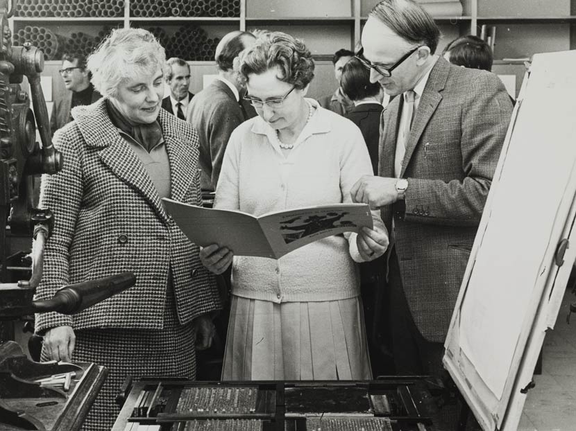 Two women and a man looking at an open book in front of an old-fashioned printing press. 
