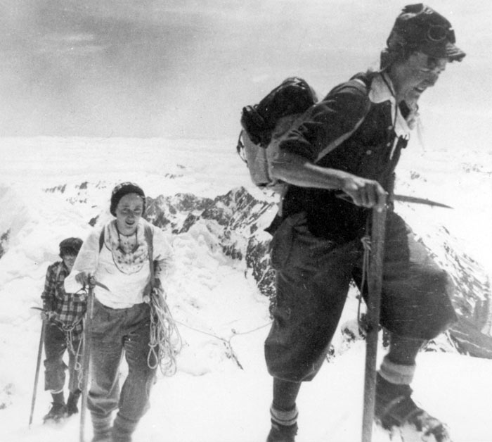 Three women, holding ropes and ice-axes, climbing up a snowy alpine ridge.