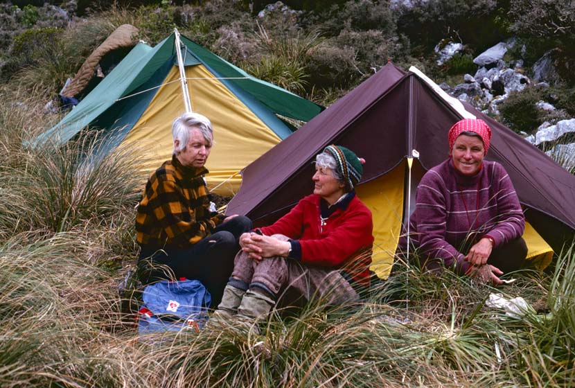 Three older women seated in an alpine setting beside two tents.