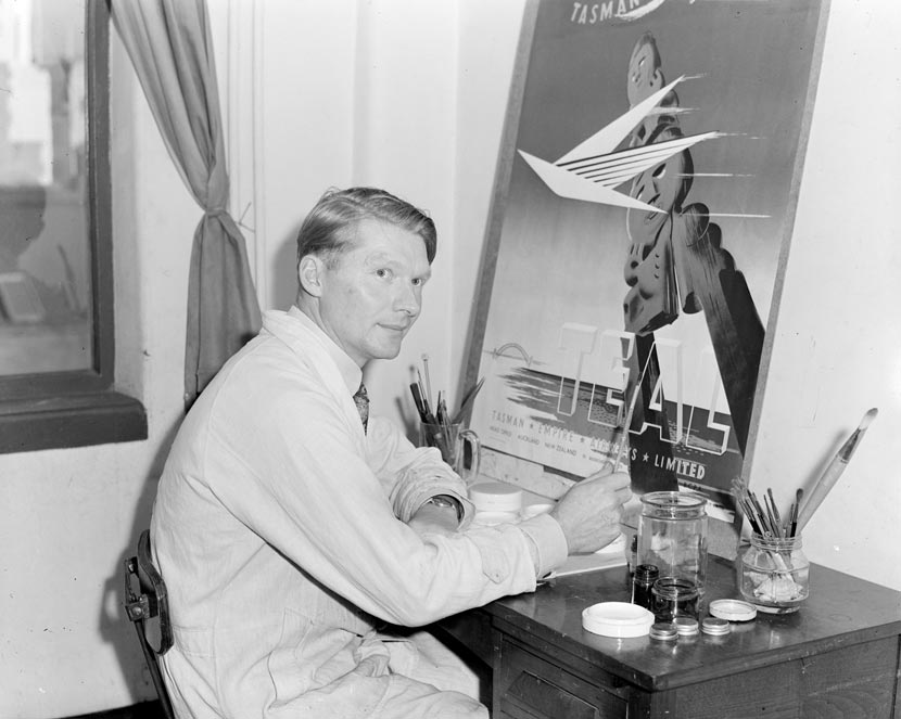 A man in a white coat is seated at a desk in front of an artwork, with paints and paintbrushes on the desk beside him.