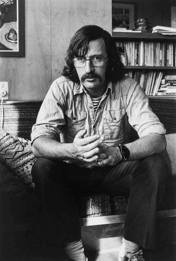 Michael King as a young man, with long hair and a moustache, seated on a sofa and looking serious.