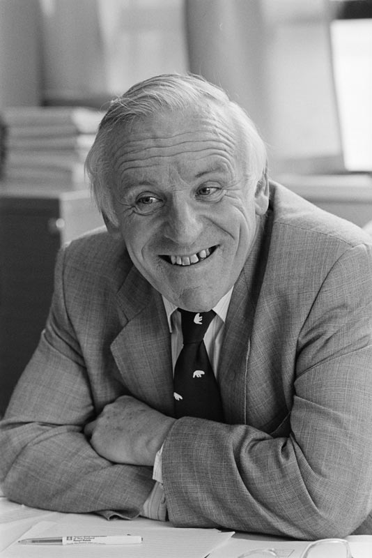 A photograph of a seated Bing Lucas leaning on a table and grinning. He wears a suit and tie.