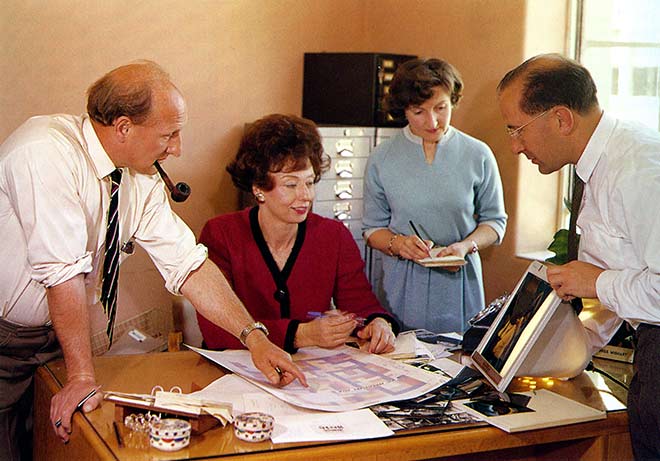 A colour photo showing Wishart seated at a desk, surrounded by three standing people. Staines points to a chart on Wishart’s desk, while Wilson shows Wishart a photograph and Lee takes notes. 