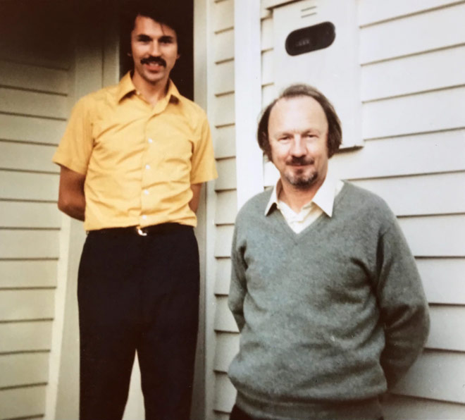 A photograph of two men standing against the outside wall of a house. Don, on the left, is in his mid-30s, slim and wearing a moustache. Bill, on the right, is middle-aged with longish hair and a goatee beard. Both men are smiling. 