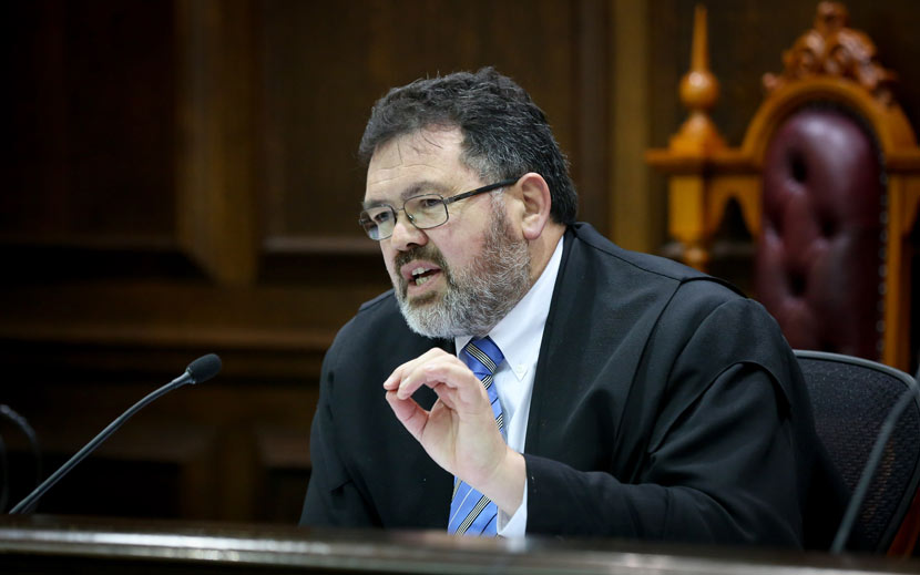 Judge Joe Williams sitting behind microphone. He has a beard and glasses and is dressed in a black legal gown, white shirt and tie.