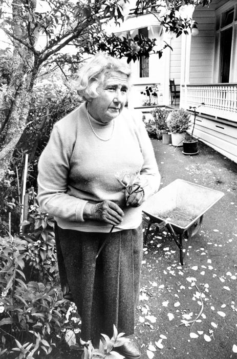 Joan Dingley standing in her front yard, holding a cut flower, with her house visible to the right.