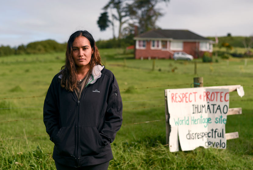 Pania Newton standing in green field with protest sign and house in the background.