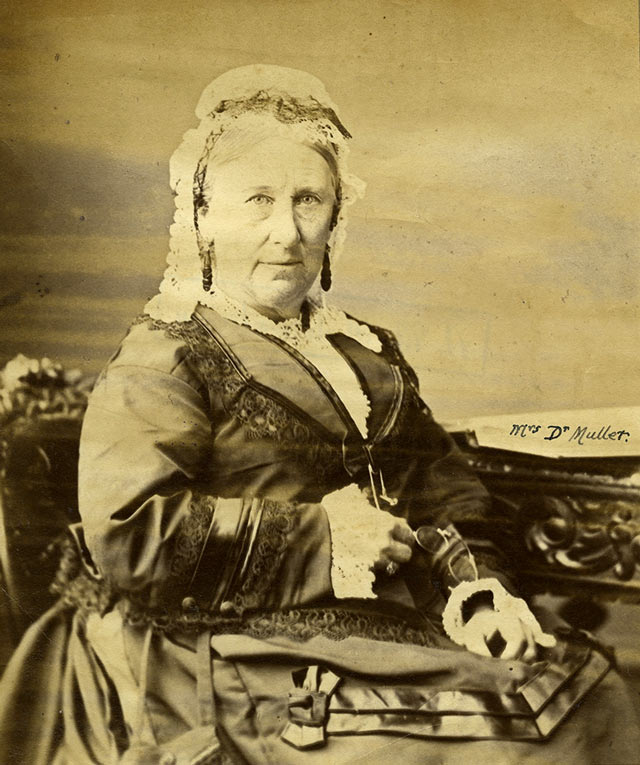 A seated studio portrait of Mary Muller looking directly into the camera. She is wearing a black dress with white lace and holding a pair of glasses in her lap.