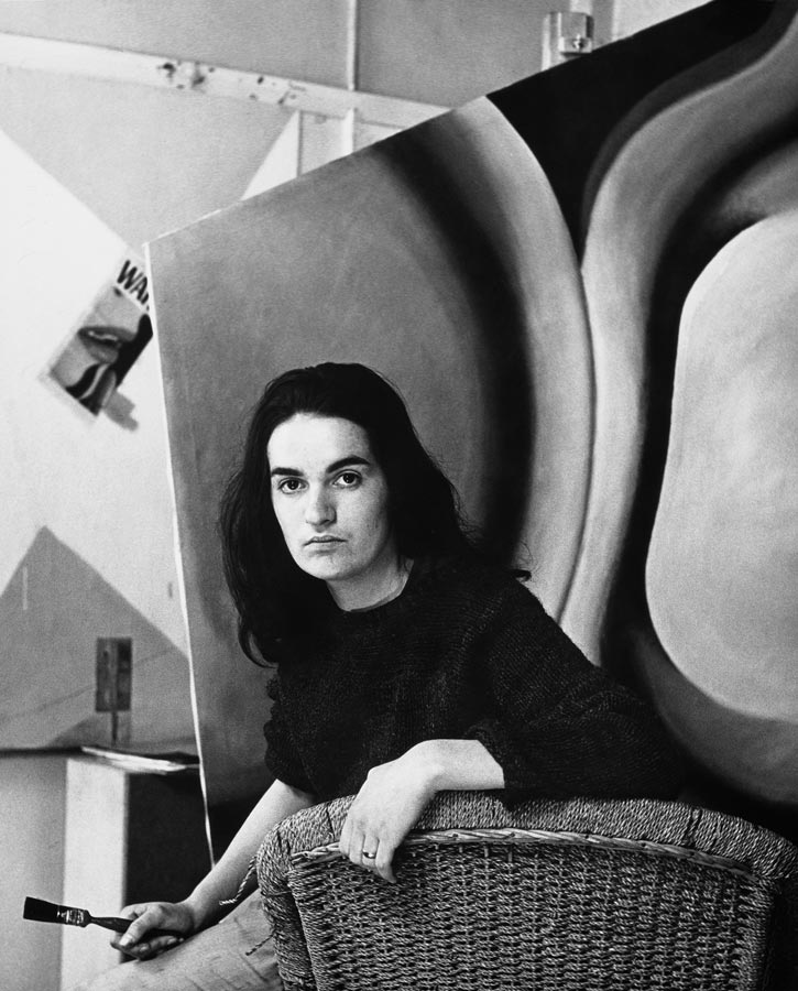 Black and white portrait of Pauline Thompson in her twenties, in front of a large abstract painting, and holding a paint brush.