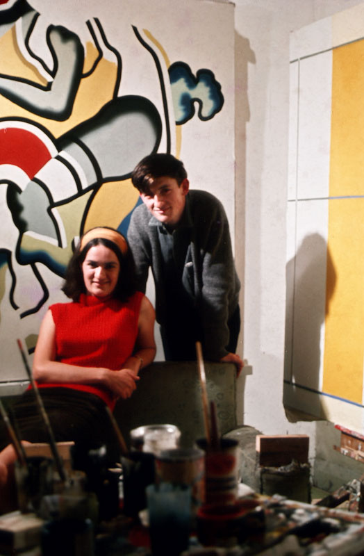 A young man and woman pose in front of a large, brightly coloured painting in yellow and red. There are paint pots and brushes in the foreground and another yellow and grey painting on the wall to the right of the viewer.