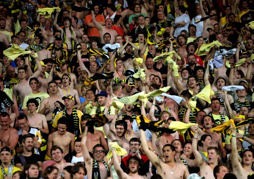 Crowd of people, many bare chested, waving black and yellow shirts and flags.
