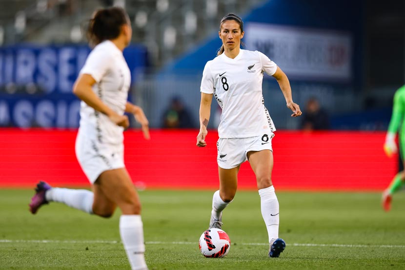 A woman in white New Zealand football uniform in the centre of photo during a football game, with a ball at her feet. A team mate runs nearby, with her back to the camera. 