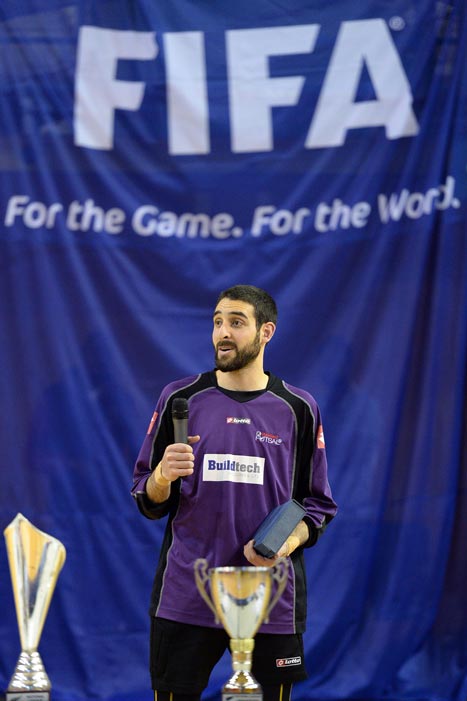 Man in purple shirt standing behind sports trophies and holding a microphone.
