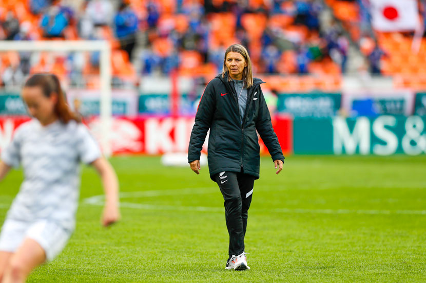 A woman in black coat with a white fern symbol walks on a football field, with a player out of focus in the foreground.